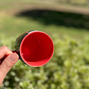 K&ouml;nnte beinhalten: Eine schwarze Kaffeetasse mit roter Innenseite wird vor einem verschwommenen gr&uuml;nen Hintergrund gehalten. Der Griff der Tasse ist rot, und der Rand hat ein schwarz-gelbes Design. Die Tasse ist leer und scheint aus Keramik zu sein.