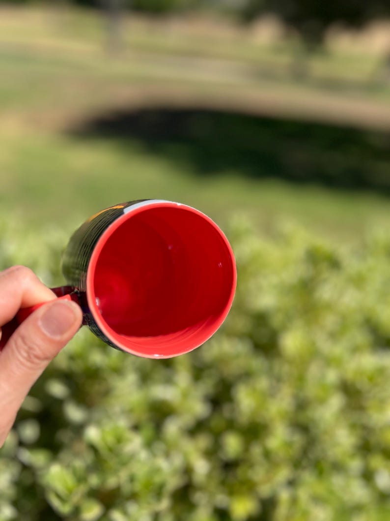 K&ouml;nnte beinhalten: Eine schwarze Kaffeetasse mit roter Innenseite wird vor einem verschwommenen gr&uuml;nen Hintergrund gehalten. Der Griff der Tasse ist rot, und der Rand hat ein schwarz-gelbes Design. Die Tasse ist leer und scheint aus Keramik zu sein.