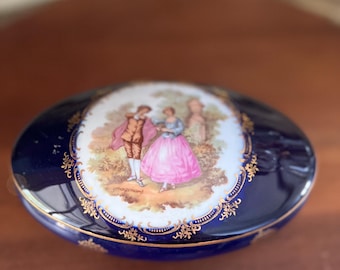 May include: An oval-shaped trinket box with a cobalt blue background and gold trim. The lid features a hand-painted scene of a couple in a garden setting.