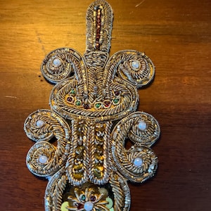 May include: A gold embroidered applique with a floral design. The applique is decorated with pearls, sequins, and beads in shades of green, red, and white.