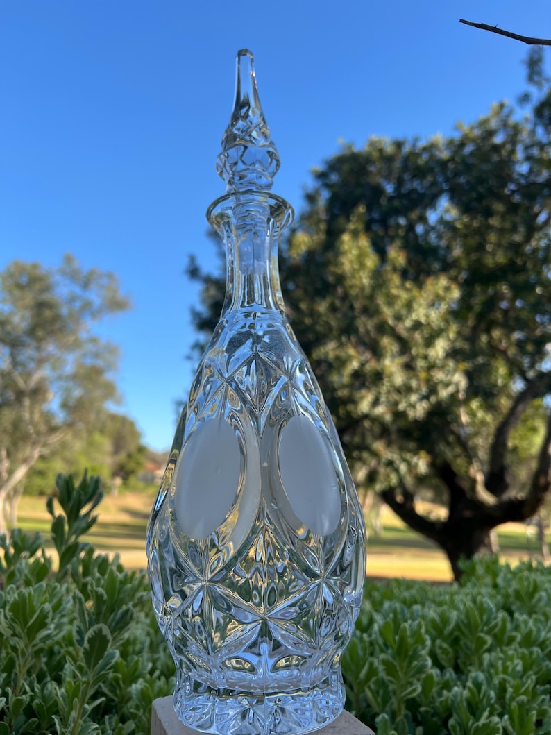 May include: A clear glass decanter with a diamond-cut pattern and a pointed stopper. The decanter has a bulbous body with two oval frosted sections. The background features green foliage and a blue sky.