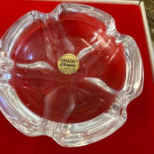 May include: A clear glass ashtray with a scalloped edge. The ashtray is sitting on a red velvet surface. The label on the ashtray reads "Cristal d'Arques France,  Genuine Lead Crystal".