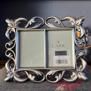 May include: A silver-colored, double-aperture picture frame with an ornate, floral design. The frame is made of pewter and features the text "CARR Frames" and a product label. The frame is resting on a patterned surface.