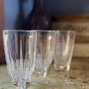 May include: Four clear, cut-glass tumblers with a detailed pattern. The glasses are arranged on a light green surface, with a blurred background featuring a dark purple vase and a wooden box.