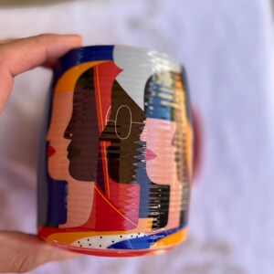 K&ouml;nnte beinhalten: Eine Keramik-Tasse mit einem farbenfrohen Design von stilisierten Gesichtern im Profil. Das Design der Tasse umfasst Rot-, Blau-, Gelb- und Schwarzt&ouml;ne. Die Tasse hat einen roten Henkel und einen wei&szlig;en Innenraum. Die Tasse wird von einer Hand gehalten.