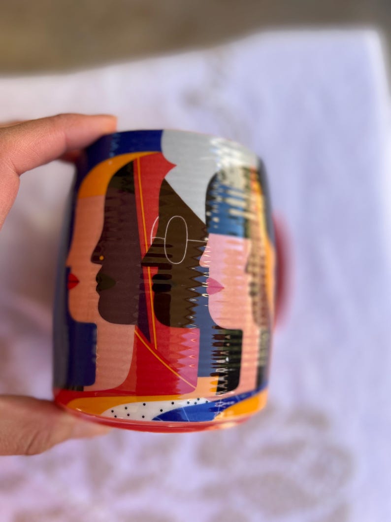 K&ouml;nnte beinhalten: Eine Keramik-Tasse mit einem farbenfrohen Design von stilisierten Gesichtern im Profil. Das Design der Tasse umfasst Rot-, Blau-, Gelb- und Schwarzt&ouml;ne. Die Tasse hat einen roten Henkel und einen wei&szlig;en Innenraum. Die Tasse wird von einer Hand gehalten.