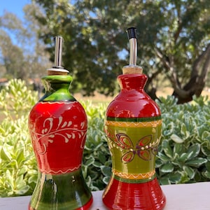 May include: Two decorative ceramic oil bottles with stainless steel spouts. One bottle is red and green with a floral design, the other is red, green, and yellow with a butterfly design. Both have cork stoppers.