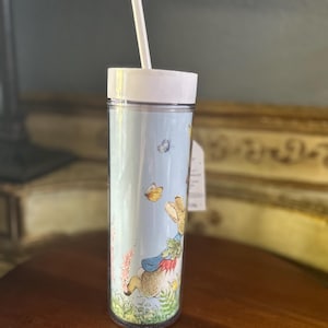 May include: A clear tumbler with a white lid and straw, featuring a whimsical illustration of a rabbit, butterflies, and flowers on a light blue background. The tumbler is cylindrical and appears to be made of plastic.