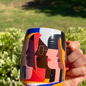 K&ouml;nnte beinhalten: Eine Keramiktasse mit rotem Henkel und einem farbenfrohen Design mit stilisierten Profilen verschiedener Gesichter. Das Design der Tasse enth&auml;lt Rot, Blau, Gelb und Schwarz und erzeugt eine lebendige und k&uuml;nstlerische &Auml;sthetik.