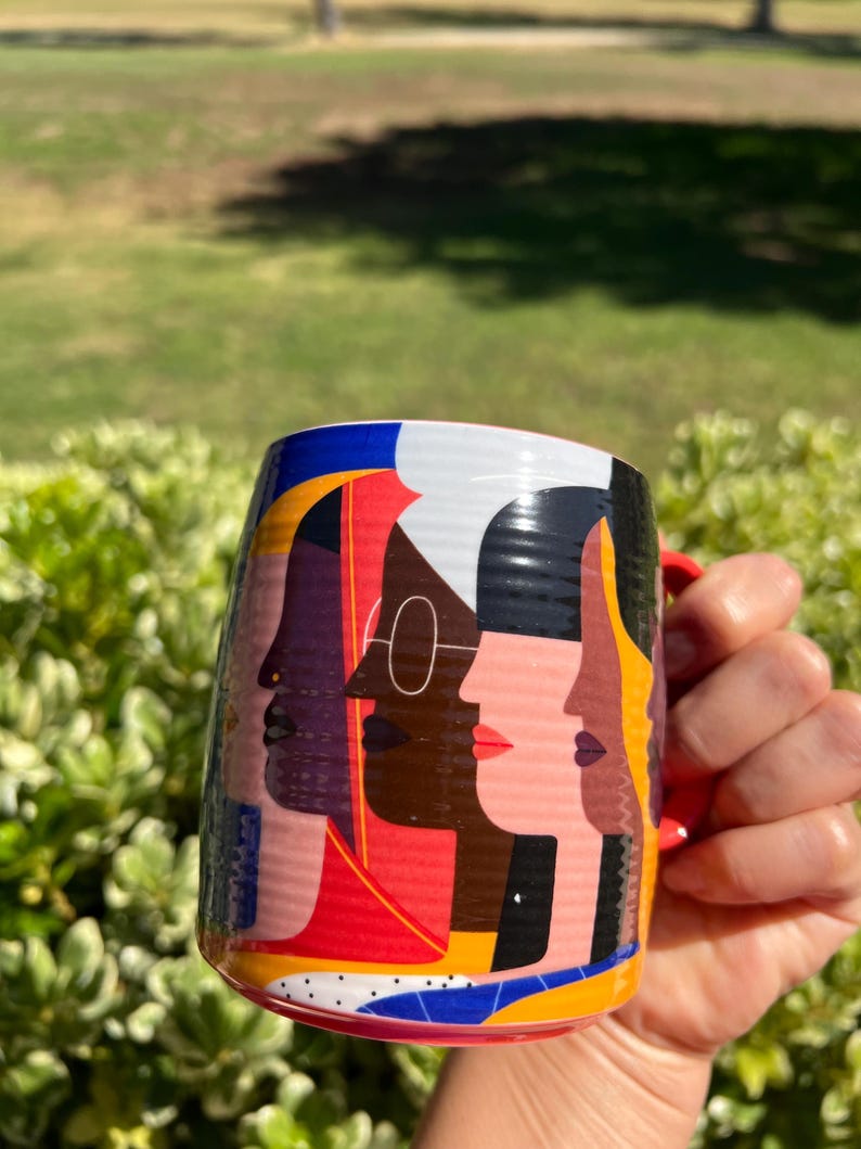 K&ouml;nnte beinhalten: Eine Keramiktasse mit rotem Henkel und einem farbenfrohen Design mit stilisierten Profilen verschiedener Gesichter. Das Design der Tasse enth&auml;lt Rot, Blau, Gelb und Schwarz und erzeugt eine lebendige und k&uuml;nstlerische &Auml;sthetik.