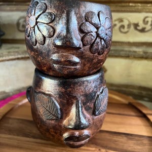 May include: Two stacked dark brown wooden cups carved into human face shapes. The top cup features flower-shaped eyes, while the bottom cup has leaf-shaped eyes. Both cups have a prominent nose, defined lips, and a textured, weathered finish.