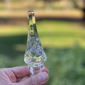 May include: A clear glass bottle stopper with a diamond-cut design on the top portion. The stopper has a long, slender shape with a cork at the top. The background is blurred, showing green foliage.