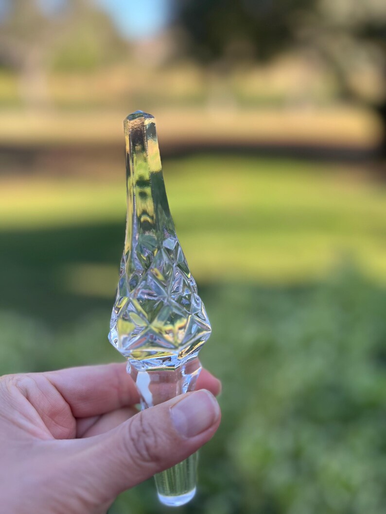 May include: A clear glass bottle stopper with a diamond-cut design on the top portion. The stopper has a long, slender shape with a cork at the top. The background is blurred, showing green foliage.