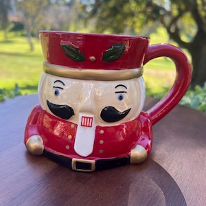 May include: A ceramic mug shaped like a toy soldier, featuring a red hat and jacket, a tan face, and black mustache. The mug has a red handle and a black belt with a gold buckle. The mug is sitting on a wooden surface.
