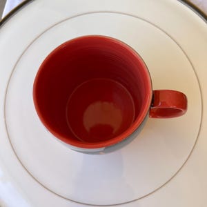 K&ouml;nnte beinhalten: Eine rote Keramik-Tasse mit passendem Henkel, auf einem wei&szlig;en Teller mit gold-schwarzem Rand. Das Innere der Tasse ist leuchtend rot und kontrastiert mit den schwarz-grauen Streifen am Rand.