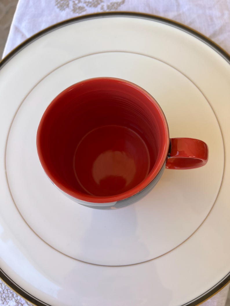 K&ouml;nnte beinhalten: Eine rote Keramik-Tasse mit passendem Henkel, auf einem wei&szlig;en Teller mit gold-schwarzem Rand. Das Innere der Tasse ist leuchtend rot und kontrastiert mit den schwarz-grauen Streifen am Rand.