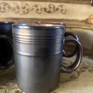 May include: A set of cylindrical ceramic mugs with a metallic dark charcoal finish. Each mug features a smooth, straight-sided body with three horizontal ridges near the top rim and a curved handle on the side. The interior of the mugs is a warm copper.