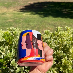 K&ouml;nnte beinhalten: Eine zylindrische Keramik-Tasse mit einem farbenfrohen Design von stilisierten weiblichen Gesichtern im Profil. Die Tasse hat einen blauen Hintergrund mit roten, gelben und wei&szlig;en Akzenten. Die Tasse wird in einer Hand vor einem Hintergrund aus gr&uuml;nem Laub und Gras gehalten.