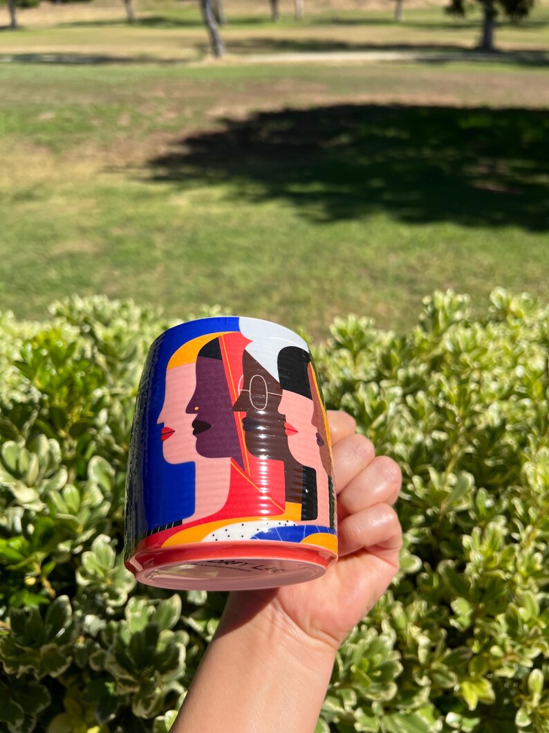 K&ouml;nnte beinhalten: Eine zylindrische Keramik-Tasse mit einem farbenfrohen Design von stilisierten weiblichen Gesichtern im Profil. Die Tasse hat einen blauen Hintergrund mit roten, gelben und wei&szlig;en Akzenten. Die Tasse wird in einer Hand vor einem Hintergrund aus gr&uuml;nem Laub und Gras gehalten.
