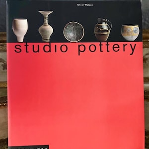 May include: A red book with the title "studio pottery" by Oliver Watson. The book cover features five ceramic pieces: a white cup, a brown vase, a brown plate, a brown jug, and a brown bowl.