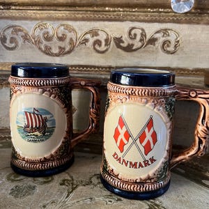 May include: Two brown ceramic beer steins with blue trim. One stein features a red and white Danish flag and the text "DANMARK". The other stein features a red and white Viking ship with the text "DANMARK".