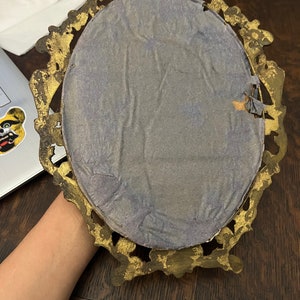 Puede incluir: Un marco de fotos ovalado de color dorado con un dise&ntilde;o decorativo. La parte posterior del marco est&aacute; cubierta con papel azul claro.
