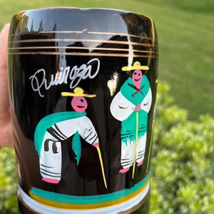 May include: Black ceramic mug with gold trim and the word "Quinga" in white script. The mug features colorful illustrations of two figures in traditional attire, with hats and staffs.