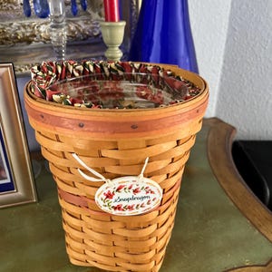 May include: Woven basket with a decorative floral tag that reads "Snapdragon". The basket has a red and brown rim and a patterned fabric lining the inside. The basket is made of light brown woven material.