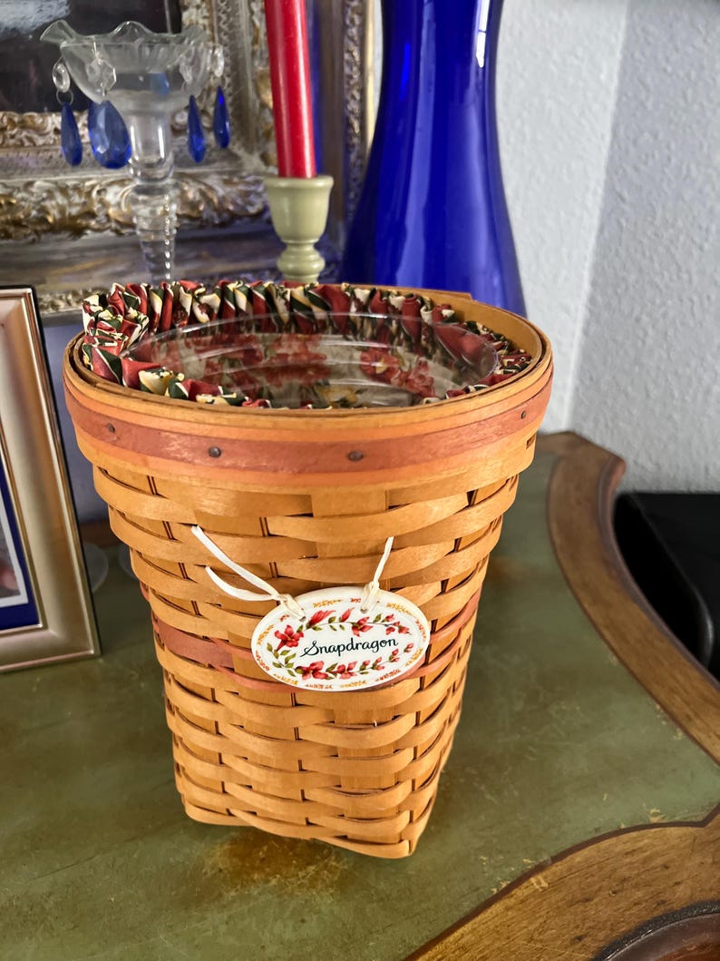 May include: Woven basket with a decorative floral tag that reads "Snapdragon". The basket has a red and brown rim and a patterned fabric lining the inside. The basket is made of light brown woven material.