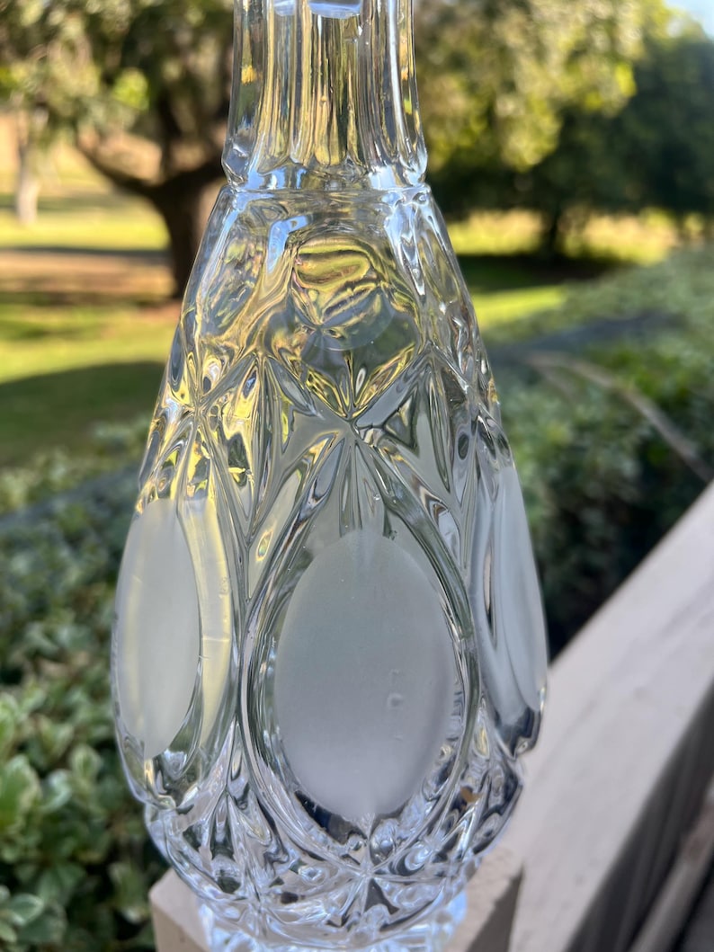 May include: A clear glass bottle with a diamond pattern and frosted oval accents. The bottle has a long neck and a decorative design, suitable for displaying liquids. The background is blurred, showing greenery.
