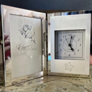 May include: Silver-toned picture frame with a clock on the right side. The clock face has black hands and black numbers. The picture frame has a white background with the word "Elegance" printed in black. The clock face has a small star with a bear inside.