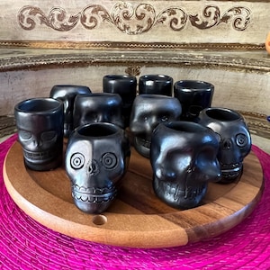 Puede incluir: Una colección de diez recipientes de cerámica negra pulida con forma de calavera, dispuestos sobre una bandeja circular de madera. Cada recipiente presenta detalles faciales tallados, incluyendo cuencas oculares, fosas nasales y dientes.