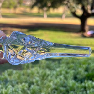 May include: A clear, faceted glass bottle stopper with a diamond pattern on the bulbous end. The stopper has a long, slender neck and is held against a blurred green background.