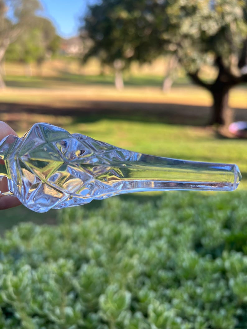 May include: A clear, faceted glass bottle stopper with a diamond pattern on the bulbous end. The stopper has a long, slender neck and is held against a blurred green background.