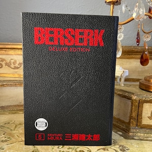 Könnte beinhalten: Ein schwarzes Hardcover-Buch mit dem Titel Berserk Deluxe Edition in roter Schrift. Der Einband zeigt einen Parental Advisory-Aufkleber, die Zahl 5 sowie den Namen des Autors Kentaro Miura in englischen und japanischen Schriftzeichen.