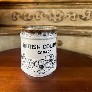 May include: White ceramic mug with a brown rim and a black floral design. The mug says "British Columbia Canada" in black lettering.