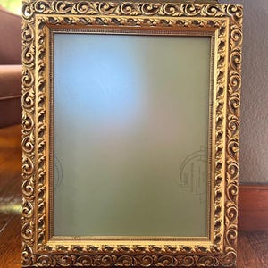 May include: Ornate gold picture frame with detailed scrollwork and a rectangular opening. The frame has a vintage aesthetic and is likely made of wood or a similar material. The frame is empty, with a light green background.