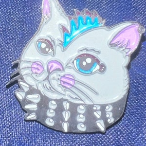 May include: A gray cat with a blue and purple mohawk, wearing a black studded collar. The cat has blue eyes and pink ears.