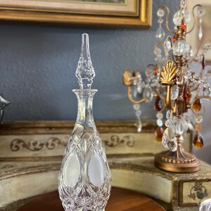 May include: A clear, cut-glass decanter with a tall, pointed stopper. The bottle has a teardrop shape with oval frosted panels and diamond-cut detailing. The decanter sits on a dark wooden surface, with a decorative chandelier in the background.