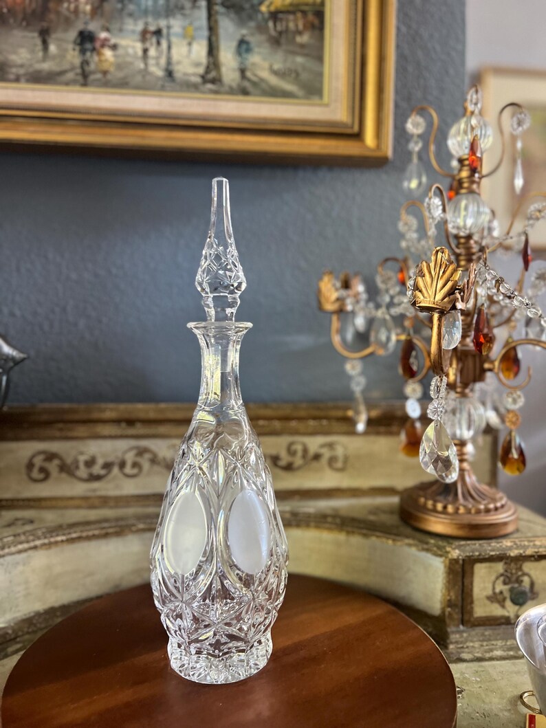 May include: A clear, cut-glass decanter with a tall, pointed stopper. The bottle has a teardrop shape with oval frosted panels and diamond-cut detailing. The decanter sits on a dark wooden surface, with a decorative chandelier in the background.