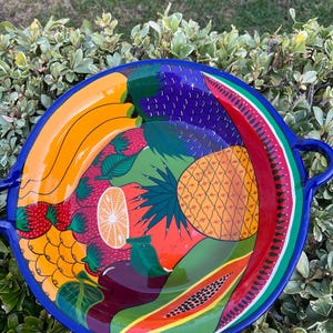 May include: A vibrant, hand-painted ceramic serving platter with a blue rim and handles. The platter is decorated with colorful illustrations of various fruits, including bananas, strawberries, pineapple, watermelon, and papaya. The design is set against a green background.