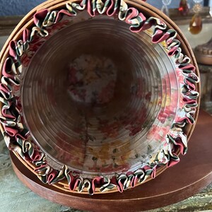 May include: A cylindrical, vintage-style hat box made of woven wood. The interior is lined with floral fabric, and the rim is trimmed with ruffled fabric in shades of red, green, and cream. The box sits on a dark wooden base.