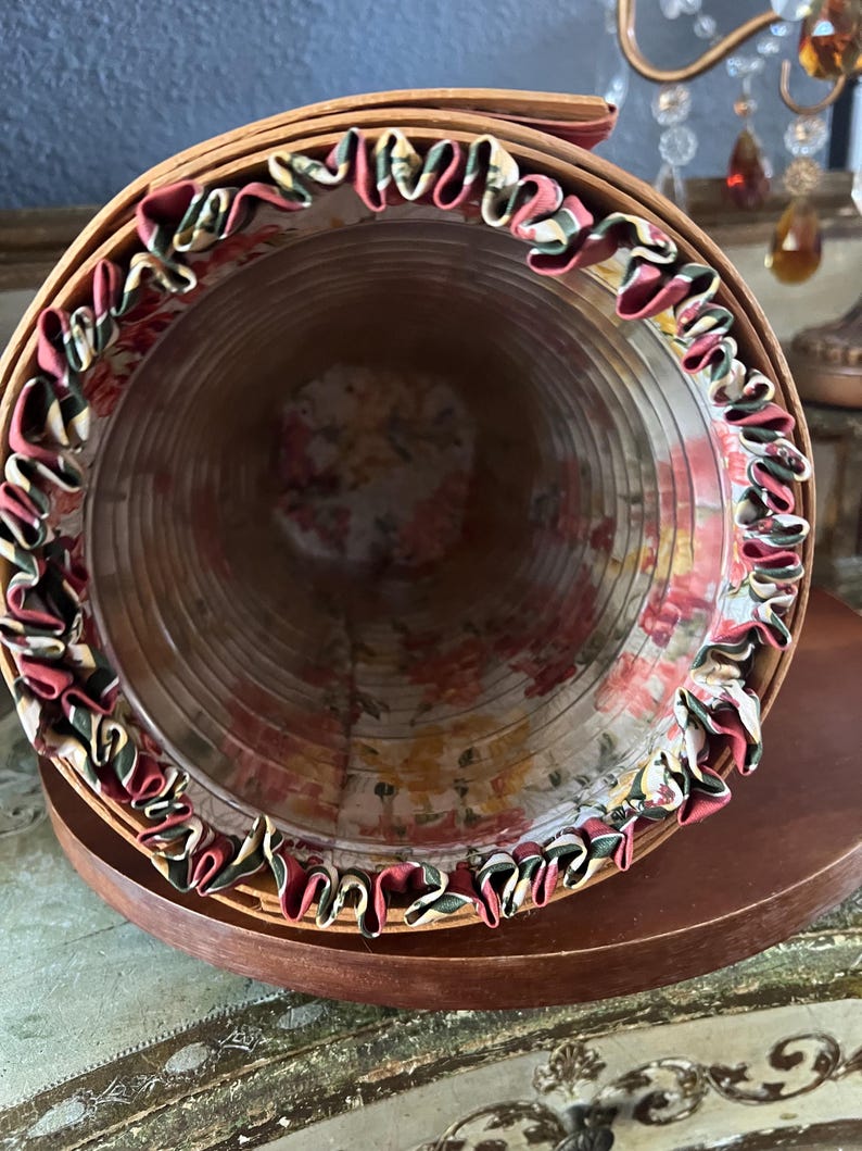 May include: A cylindrical, vintage-style hat box made of woven wood. The interior is lined with floral fabric, and the rim is trimmed with ruffled fabric in shades of red, green, and cream. The box sits on a dark wooden base.