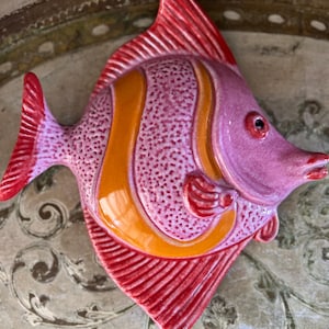 May include: A ceramic fish wall ornament featuring a textured pink body with smooth orange vertical stripes and vibrant red fins. The fish has a glossy finish, a small pectoral fin, and a distinct eye, showcasing a stylized aquatic design.
