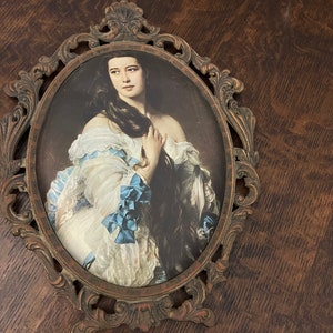 Puede incluir: Un retrato ovalado enmarcado de una mujer con el pelo largo y oscuro que lleva un vestido blanco con detalles azules. El marco es ornamentado y est&aacute; hecho de metal con un acabado marr&oacute;n.