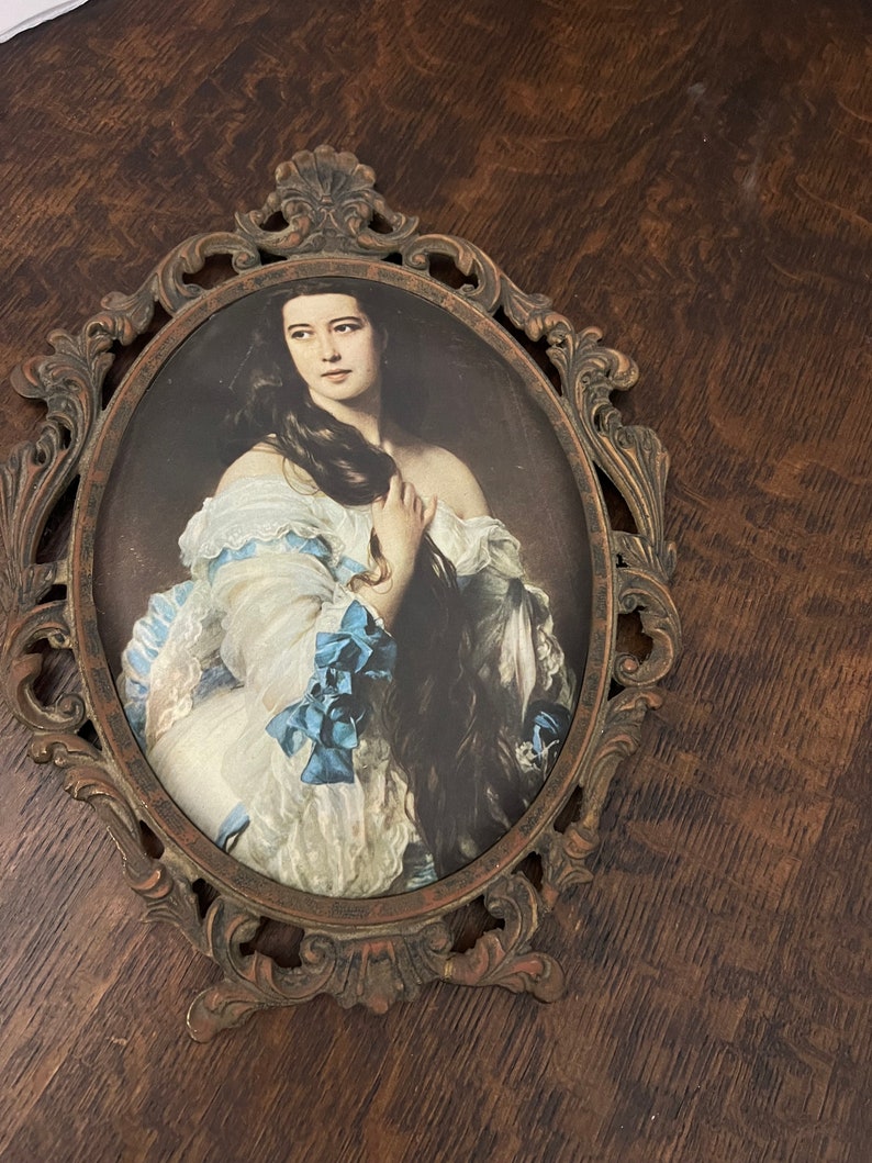 Puede incluir: Un retrato ovalado enmarcado de una mujer con el pelo largo y oscuro que lleva un vestido blanco con detalles azules. El marco es ornamentado y est&aacute; hecho de metal con un acabado marr&oacute;n.