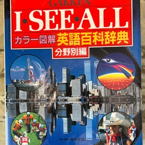 Op de afbeelding: Een rode, blauwe en witte boekomslag met de titel "I SEE ALL" in grote witte letters. Het boek is een Engelstalige encyclopedie in de Japanse taal met illustraties van een zeilboot, een skyline, een vliegtuig, een voetbalwedstrijd en een meisje in een cowboyhoed.