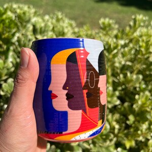 K&ouml;nnte beinhalten: Eine zylindrische Keramiktasse mit blauem Hintergrund und einer farbenfrohen Illustration verschiedener Gesichter. Die Tasse wird von einer Hand vor einem Hintergrund aus gr&uuml;nem Laub gehalten.