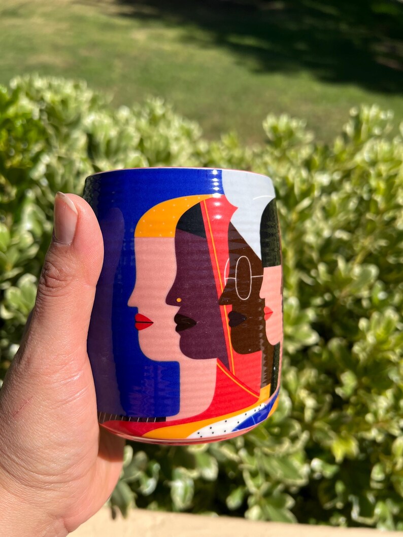 K&ouml;nnte beinhalten: Eine zylindrische Keramiktasse mit blauem Hintergrund und einer farbenfrohen Illustration verschiedener Gesichter. Die Tasse wird von einer Hand vor einem Hintergrund aus gr&uuml;nem Laub gehalten.