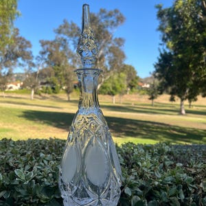 May include: A clear glass decanter with a diamond-cut pattern and a frosted oval design. The stopper is a tall, pointed shape with a diamond pattern. The decanter is displayed outdoors with a green background.
